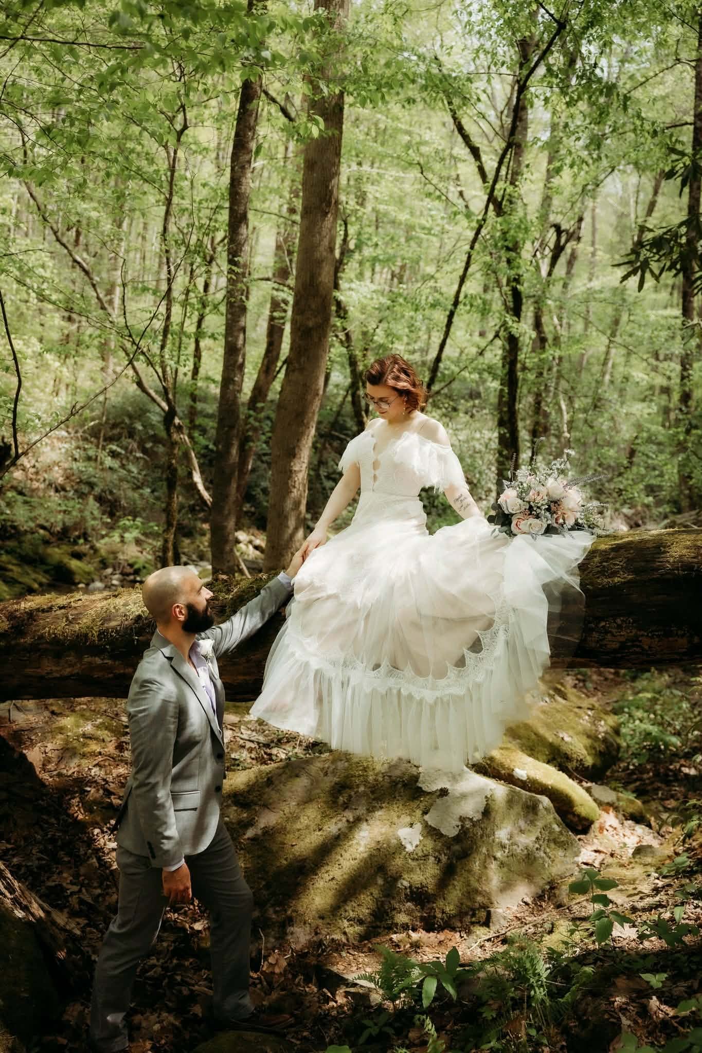 Groom helping his bride across mossy rocks in the forest